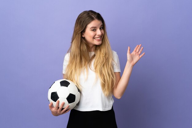 Mujer joven rubia jugador de fútbol aislada en el espacio púrpura saludando con la mano con expresión feliz