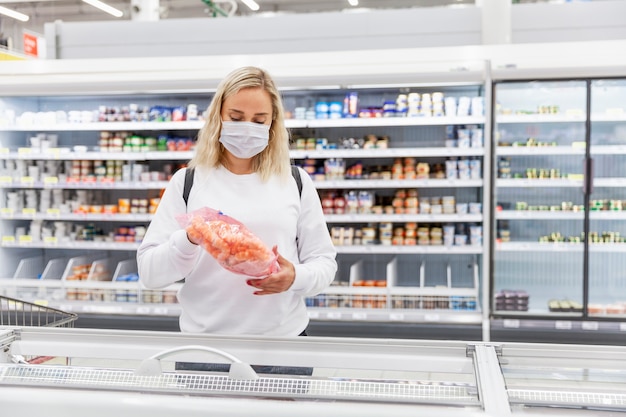Mujer joven rubia con una máscara médica en el departamento de alimentos congelados. Salud y nutrición adecuada durante una pandemia.