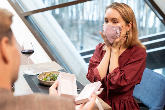 Mujer joven rubia probándose el presente de su novio y mirándolo en la cena mientras ambos están sentados junto a la mesa servida en un restaurante de lujo