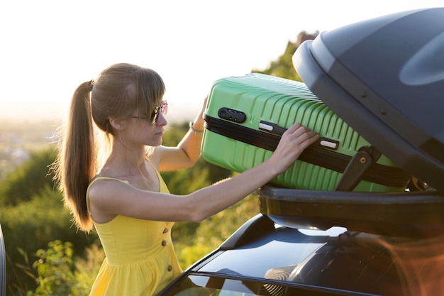 Mujer joven sacando la maleta verde de la parrilla del techo del coche. Concepto de viajes y vacaciones.