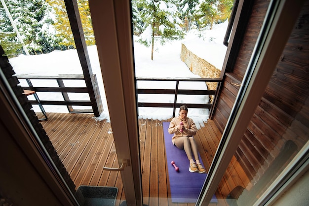 Mujer joven sana y en forma tumbada en una estera de yoga con pesas en las manos y entrenando abdominales Vista desde la ventana en la terraza Naturaleza nevada de invierno en el fondo