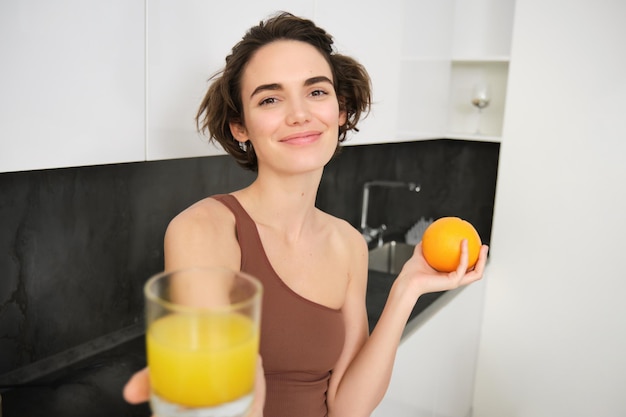 Mujer joven sana y hermosa que le da un vaso de jugo recién exprimido sosteniendo una naranja y s