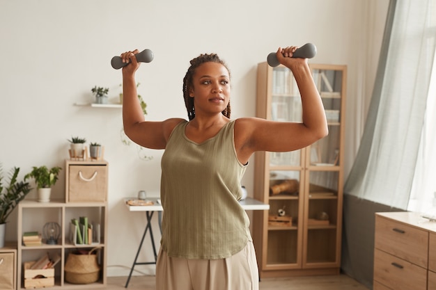 Foto mujer joven sana levantando sus manos y entrenando con pesas en la sala de estar