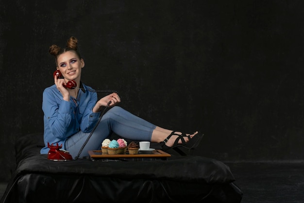 Mujer joven en sandalias de tacón habla por teléfono fijo retro Bandeja de cupcakes con fondo negro crema multicolor