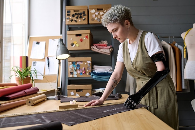 Mujer joven sastre con prótesis de brazo negro de pie junto a la mesa en el taller