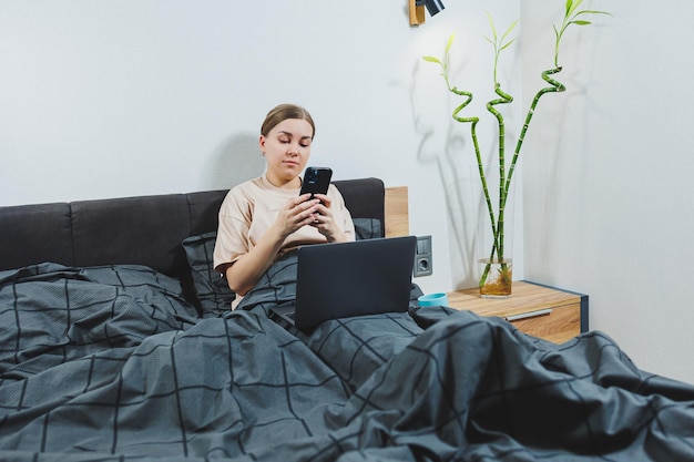 Mujer joven satisfecha con el teléfono que continúa trabajando en la computadora portátil Mujer sentada con la computadora en la cama estudiando en casa el aprendizaje a distancia y el concepto de trabajo independiente remoto