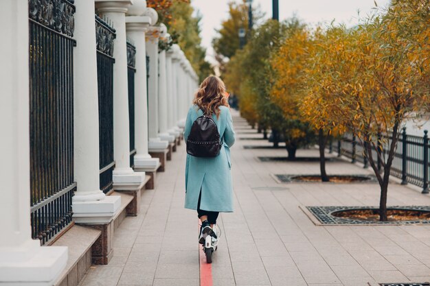 Mujer joven con scooter eléctrico en abrigo azul en el carril bici en la ciudad