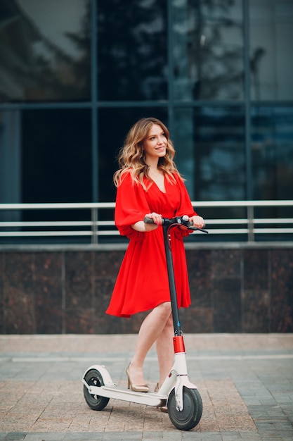 Mujer joven con scooter eléctrico en vestido rojo en la ciudad