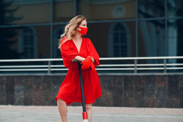 Mujer joven con scooter eléctrico en vestido rojo y guantes con mascarilla médica en la ciudad