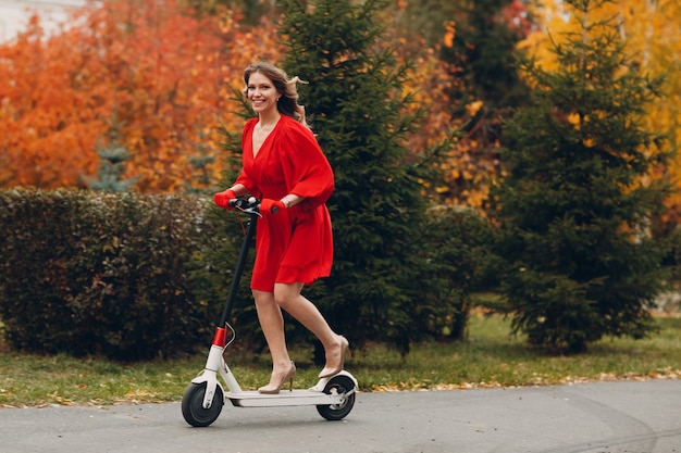 Mujer joven con scooter eléctrico en vestido rojo en el parque de la ciudad de otoño