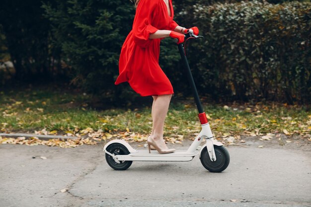 Mujer joven con scooter eléctrico en vestido rojo en el parque de la ciudad de otoño