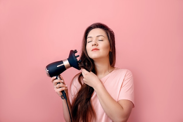 Mujer joven con secador de pelo en manos sobre fondo rosa