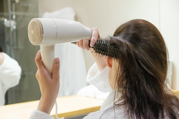 Mujer joven secar el cabello con secador de pelo y peinar el cabello con  cepillo redondo en el baño