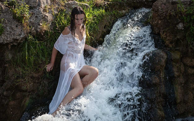 Mujer joven semidesnuda con un vestido blanco disfruta de la frescura y el frescor en los chorros de agua de la cascada