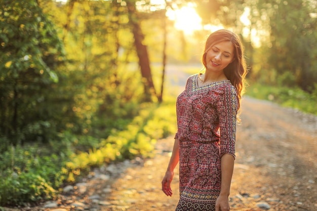 Mujer joven en el sendero del bosque, mirando hacia abajo sobre su hombro, el sol detrás de ella, la retroiluminación crea un bonito efecto bokeh en el fondo.