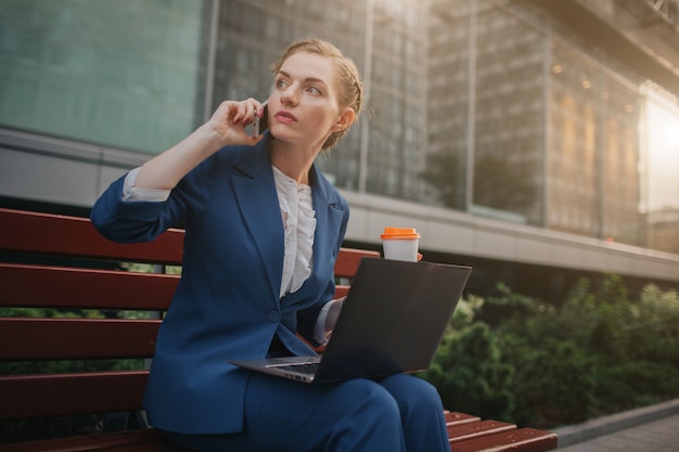 Mujer joven sentada al aire libre y usando la computadora portátil. empresaria trabajando sosteniendo la taza de café. y hablando por teléfono al mismo tiempo