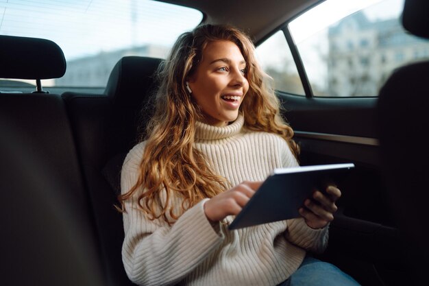 Mujer joven sentada en el asiento trasero del coche con una tableta en la mano Business technologie concierto en línea