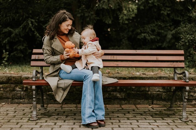 Foto mujer joven sentada en un banco con una linda niña en el parque de otoño