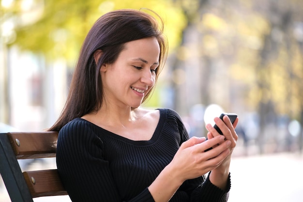 Mujer joven sentada en un banco del parque mirando su teléfono celular al aire libre en un cálido día de verano Comunicación y concepto de conexión móvil