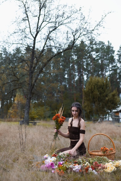 Una mujer joven sentada en el bosque y recolecta flores que yacen en el suelo con una canasta cerca.