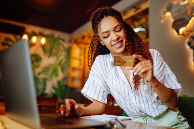 Mujer joven sentada en un café haciendo compras en línea usando una tarjeta de crédito y una computadora portátil