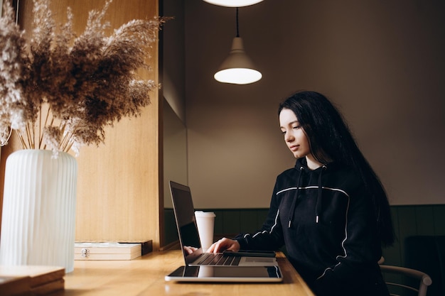 Mujer joven sentada en una cafetería en una mesa de madera tomando café y usando un teléfono inteligente En la mesa hay una computadora portátil Chica navegando por Internet chateando blogs Mujer sosteniendo el teléfono y mirando su pantalla