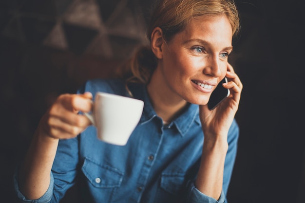 Mujer joven sentada en la cafetería, usando teléfono y bebiendo café