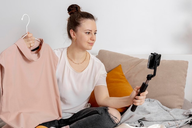 Mujer joven sentada en la cama en casa y tomando fotos de su ropa vieja para venderla en línea Vender en el comercio electrónico del sitio web Reutilizar el concepto de segunda mano Estilo de vida sostenible del consumidor consciente