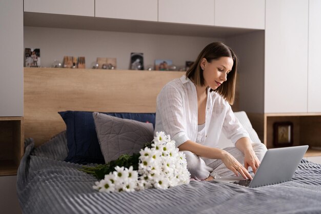 Mujer joven sentada en la cama en pijama con placer disfrutando de flores blancas charlando usando una computadora portátil