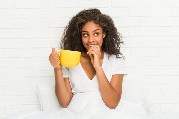 Mujer joven sentada en la cama sosteniendo una taza de café relajado pensando en algo mirando un espacio de copia.