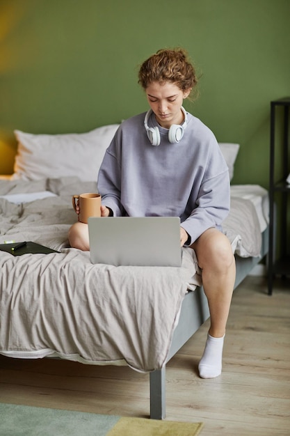 Mujer joven sentada en la cama con una taza de café y escribiendo en la computadora portátil en casa