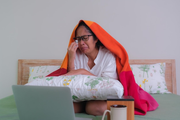 Una mujer joven sentada en la cama viendo una película en su computadora portátil llorando Una taza de té y una caja de papel tisú frente a ella