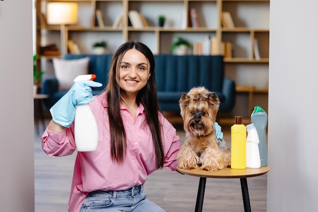 Mujer joven sentada cerca de la mesa con productos de limpieza en casa y su perrito también está sentado en la mesa