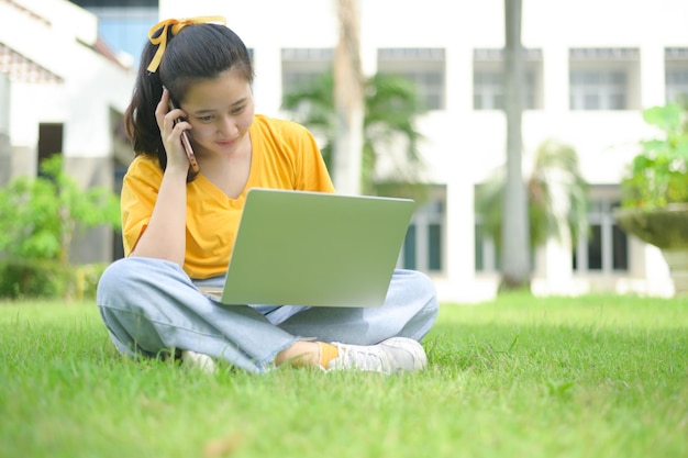 Mujer joven sentada en el césped hablando por teléfono y usando una computadora portátil