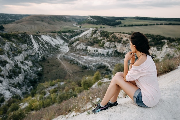 Mujer joven sentada en la cima de una montaña, mirando a un cañón de piedra blanca.