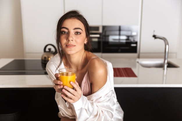 Mujer joven sentada en la cocina, sosteniendo un vaso con jugo de naranja