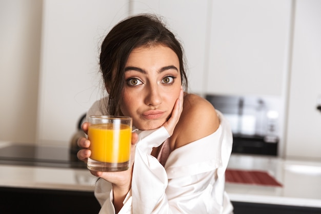 Mujer joven sentada en la cocina, sosteniendo un vaso con jugo de naranja
