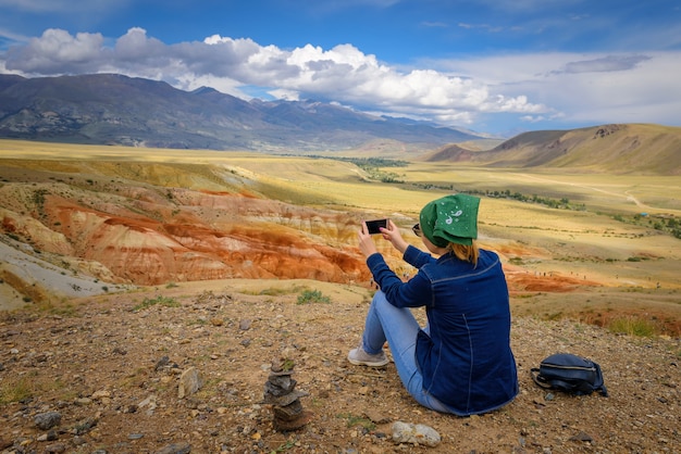 Mujer joven sentada en la colina y tomando fotos en el teléfono inteligente de un hermoso paisaje de montaña