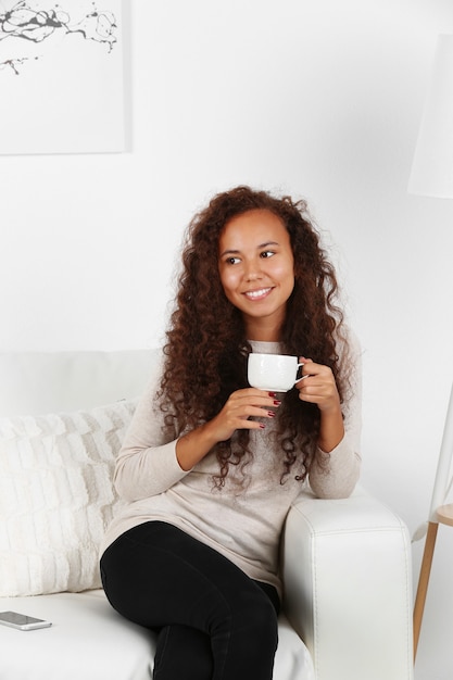 Mujer joven sentada en un cómodo sofá con una taza de café en las manos en la sala blanca