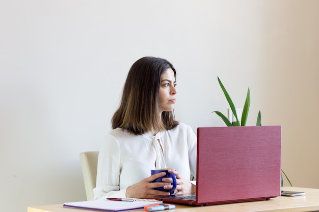 Mujer joven sentada con una computadora portátil y una taza de café trabajando desde casa