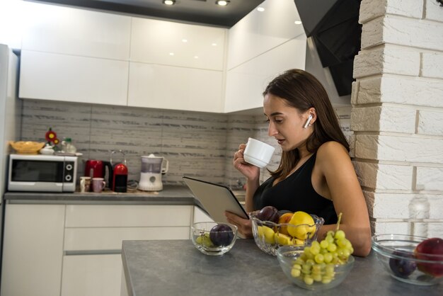 Mujer joven sentada delante de la computadora portátil abierta y tomando café en su cocina. Disfrutando el tiempo en casa