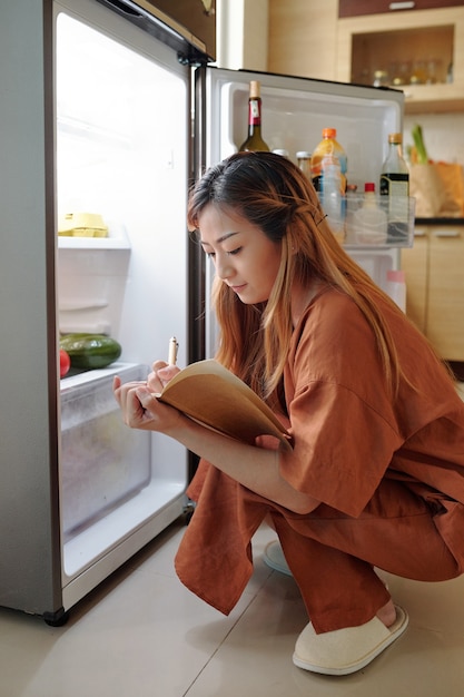 Mujer joven sentada delante de la nevera y escribiendo la lista de compras en el planificador