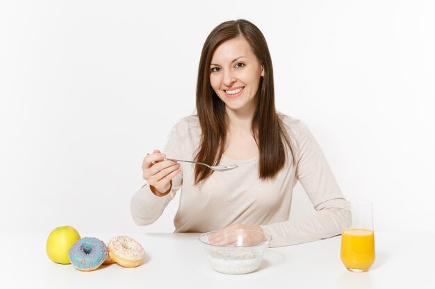 Mujer joven sentada en el desayuno de mesa con cereales y leche, jugo de naranja en vidrio, donas aislado sobre fondo blanco. Nutrición adecuada, comida deliciosa y sabrosa, estilo de vida saludable. Área para copiar espacio.