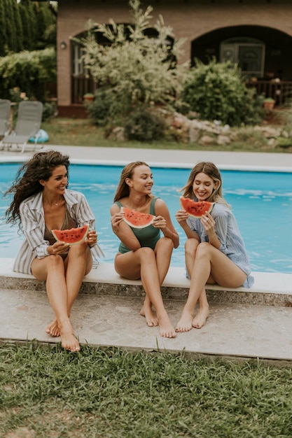 Mujer joven sentada junto a la piscina y comiendo sandía en el patio trasero de la casa