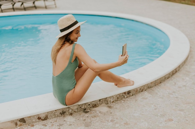 Mujer joven sentada junto a la piscina y tomando una foto selfie con teléfono móvil en el patio trasero de la casa