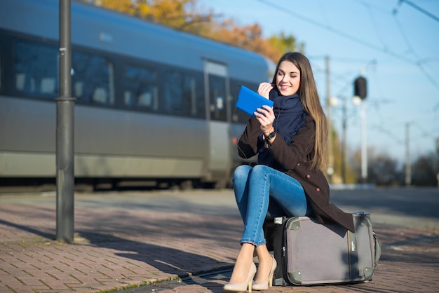 Mujer joven sentada en una maleta usando una tableta en la estación de tren