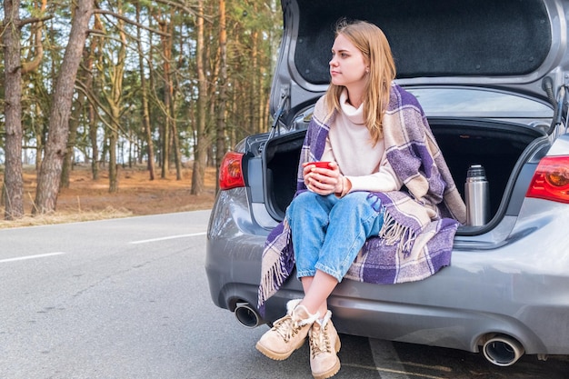 Mujer joven sentada en el maletero del coche con tazas de café y termos al lado de la carretera en el bosque