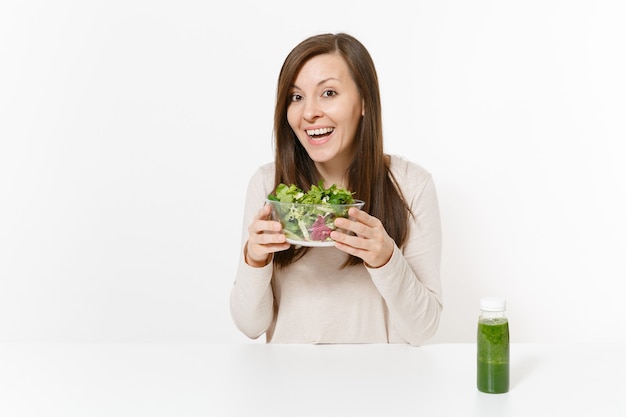 Mujer joven sentada a la mesa con batidos de desintoxicación verde, ensalada en un recipiente de vidrio aislado sobre fondo blanco. Nutrición adecuada, comida vegetariana, estilo de vida saludable, concepto de dieta. Área con espacio para copiar.