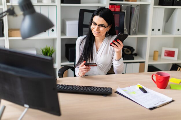 Foto una mujer joven está sentada a la mesa, sosteniendo una tarjeta bancaria y un teléfono.