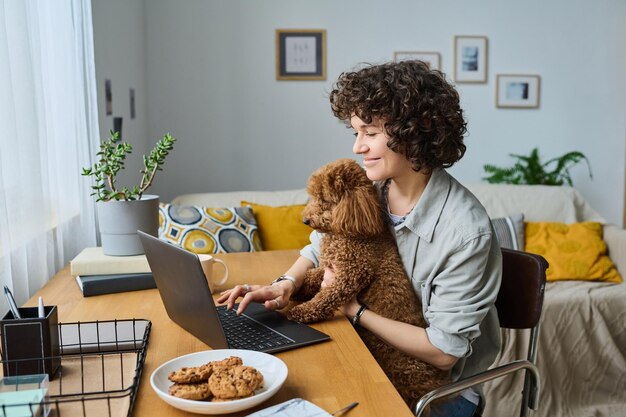 Mujer joven sentada en la mesa con su perro y usando una computadora portátil para hacer su trabajo en línea